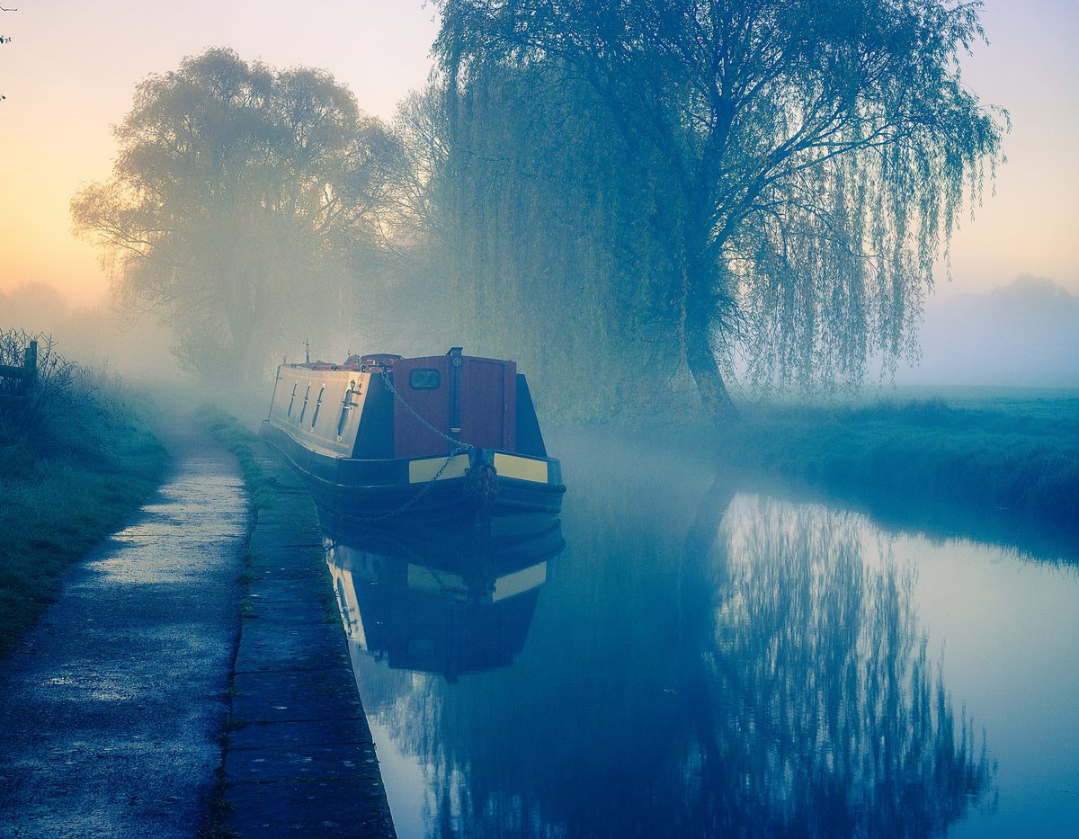 Narrowboat moored beneath a weeping willow on a misty canal at dawn